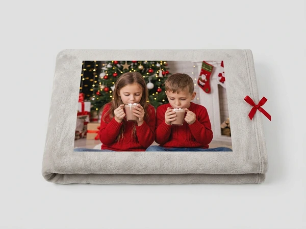 Folded plush blanket of a girl and a boy in red sweaters sipping hot cocoa by a Christmas tree