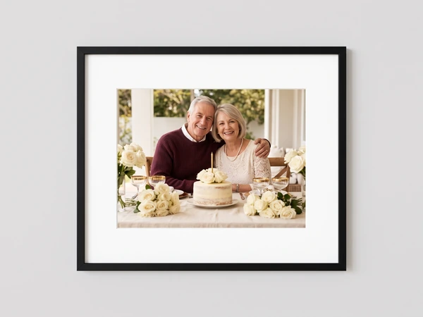 Black framed print of an older couple at a table with a celebration cake and white roses