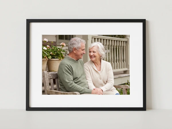 Black framed print of an elderly couple sitting on a garden bench by a porch