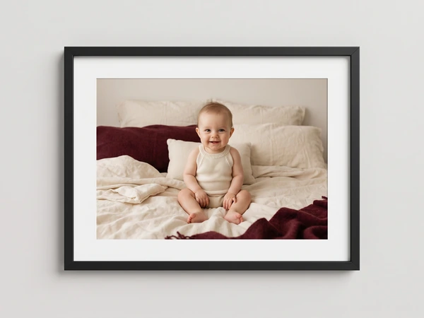 Black framed print of a smiling baby sitting on a bed with cream and burgundy blankets