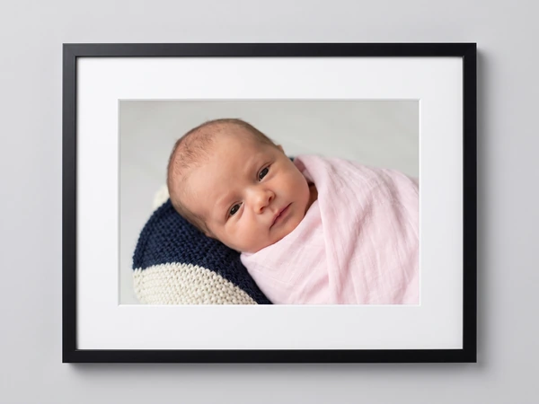 Black framed print of a newborn swaddled in pink resting on a navy and white knit pillow