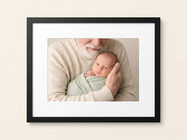 Black framed print of a bearded man cradling a newborn in a green swaddle