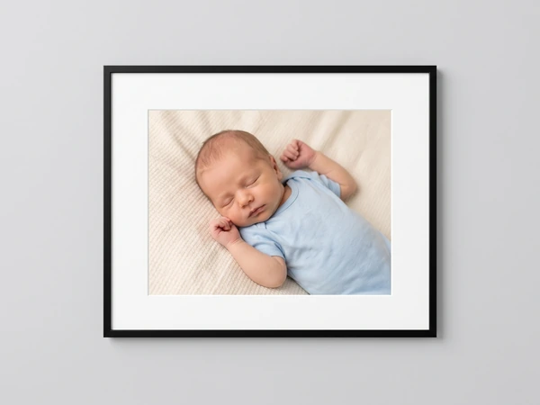Black framed print of a newborn in a blue sleepsuit stretching on a cream blanket