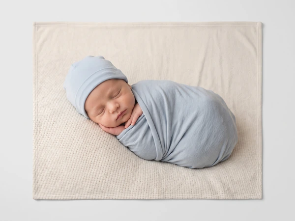 Fleece blanket printed with a newborn in a blue hat and wrap asleep on cream knit