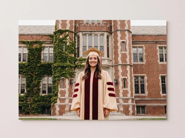 Metal print of a graduate in a cream and burgundy gown before an ivy-covered brick building