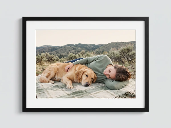 Black framed print of a woman lying beside a sleeping golden retriever on a blanket outdoors