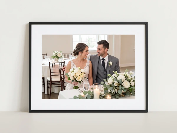 Black framed print of a bride and groom at a table set with white flowers and candles