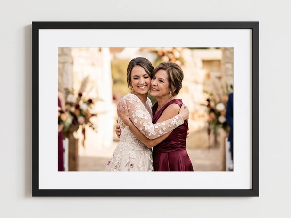Black framed print of a bride in lace hugging her mother in burgundy