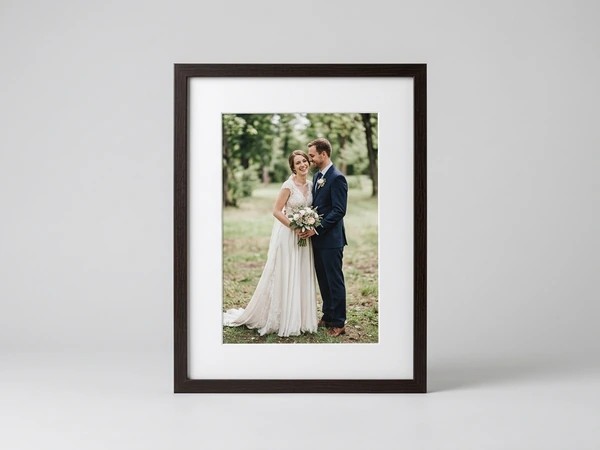 Black framed portrait print of a bride and groom standing in a wooded clearing