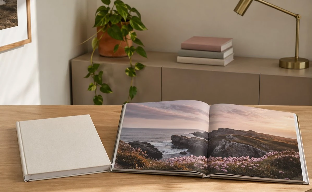 Open photo book showing a coastal landscape spread on a wooden desk, with a closed linen-bound photo book beside it