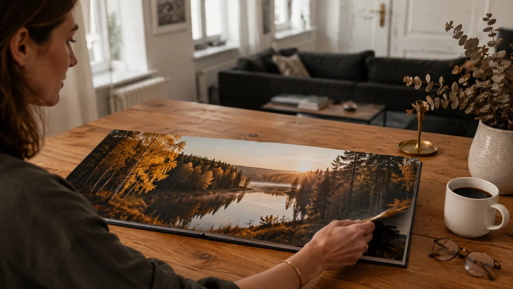 Woman viewing an open layflat photo book on a wooden table, with a forest sunset panorama spanning both pages seamlessly